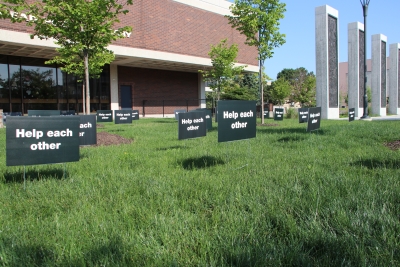 Multiple small black yard signs reading "help each other" on the library lawn