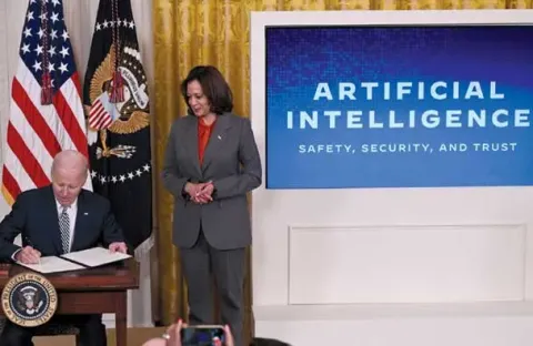Kamala Harris standing next to Biden, who is sitting at a podium. There is a slideshow in the background with the words "Artifical Intelligence" on it.
