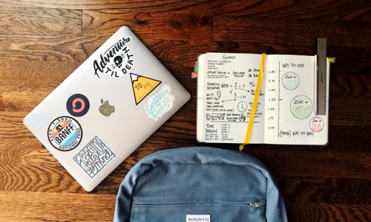 Macbook with stickers, open planner, and blue backpack on table